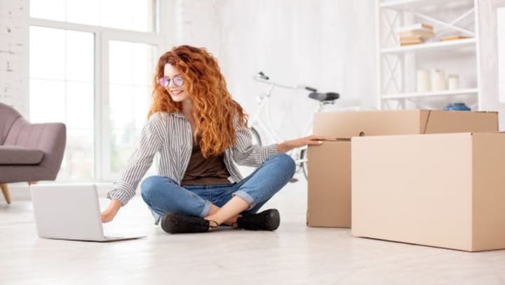 A smiling woman working on a laptop surrounded by moving boxes.