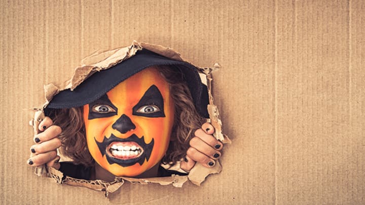 A little girl with her face painted like a pumpkin poking her head through a hole in a box.